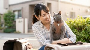 住宅街の屋外で、日本人女性が猫用のベスト型ハーネスと迷子札付きの首輪を装着した愛猫を優しく抱き寄せ、笑顔を見せている。傍らには開いたキャリーバッグがあり、安全な外出を象徴している。